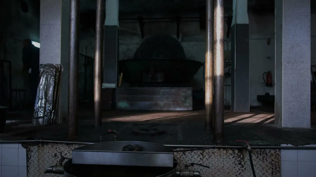 The iron press and granite wheel in the sleeping mill, lit by shafts of light.